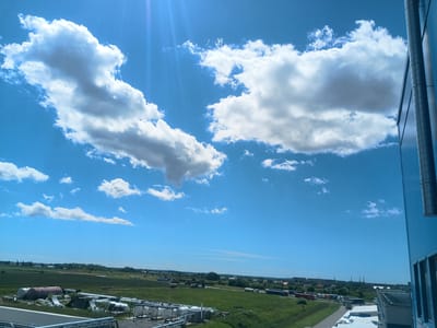 This media file depicts an outdoor scene under a bright, blue sky dotted with fluffy white clouds. The sun is high in the sky, suggesting it is midday. A large, modern building with a blue, reflective facade occupies the right side of the frame, with visible vertical piping.

Below the sky, a sprawling industrial or semi-rural landscape unfolds. In the foreground and midground, there are various metal tanks and pipes, some appearing weathered and unused, suggesting a storage or processing area. Further in the distance, a line of trucks is visible on a road, hinting at ongoing logistical activity. Beyond the immediate industrial elements, there is a verdant green field and a scattering of trees and buildings, indicating a settlement or town in the background. The overall impression is one of a working industrial area set against a backdrop of open countryside. There are no people visible in the scene, and no text is discernible. The weather appears to be clear and sunny. The location context provided is Rimkai, Lithuania.