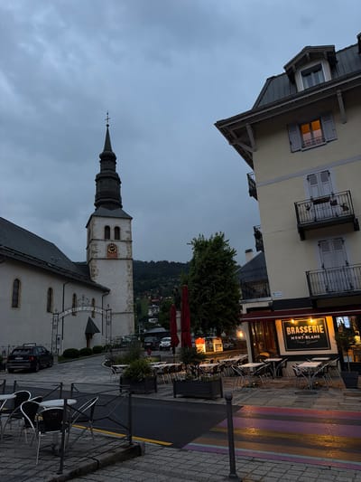 The image depicts a town square in Saint-Gervais-les-Bains, France, at dusk. A prominent church tower with a clock face stands on the left, adjacent to a white building with arched windows. An ornate archway with "JARDIN PUBLIC" inscribed on it leads to the church. In the foreground, several tables and chairs are set up outside a brasserie named "BRASSERIE du MONT-BLANC Saint-Gervais." The street and crosswalk are wet, reflecting the lights from the brasserie and streetlights. The sky is overcast, and a few cars are parked on the side of the road. The background shows a hillside covered in trees and buildings, indicating a mountainous region. There are no people visible in the image.