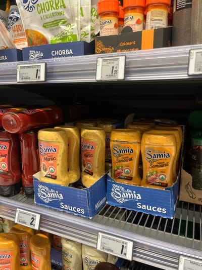 The image shows a grocery store shelf in Saint-Brice-Courcelles, France, filled with various food items. On the top shelf, there are packages of soup mix, specifically "Chorba à l'Algérienne" and "Harissa à la Marocaine." Below them are a few bottles of red food coloring labeled "Samia Colorant Alimentaire." The bottom two shelves are stocked with several bottles of "Samia Sauces" in various flavors including "Sauce Maroc," "Sauce Samourai Piments," and "Sauce Ketchup." The price tags indicate that the "Chorba" soup mix costs €1.85, the food coloring costs €1.35, and the "Samourai" sauce costs €1.98. The bottles are arranged in boxes, with "Samia Sauces" printed on the front. The shelves are made of metal and are well-lit. The image captures a typical grocery store environment.