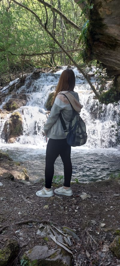 A young woman stands on a dirt path next to a waterfall at Бачковски манастир, Bulgaria. She is wearing black leggings, white sneakers, a grey hoodie with a beige section on the back, and a dark-colored backpack. The waterfall is to her left, cascading over rocks and forming a stream. Lush green trees and vegetation surround the area, with a rock formation visible in the upper left corner. The dirt path is covered with leaves, small rocks, and roots.