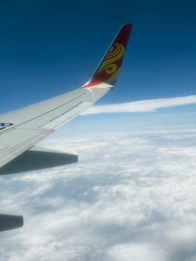 The image displays an aerial view from an airplane window, featuring a portion of the aircraft's wing against a clear blue sky and a layer of clouds below. The wing, predominantly white, exhibits a sleek design with visible panel lines and what appear to be navigation lights. At the tip of the wing, a red and yellow emblem, possibly a logo or livery, is prominent. The vast expanse of the sky is a deep blue, with wispy white clouds forming a textured layer beneath the aircraft. There are no visible landmarks or cityscapes suggesting a specific location within Changzhi City, China, as the view is entirely above the cloud cover. The scene is devoid of people or any discernible human activity, focusing solely on the airplane's structure and the atmospheric environment.