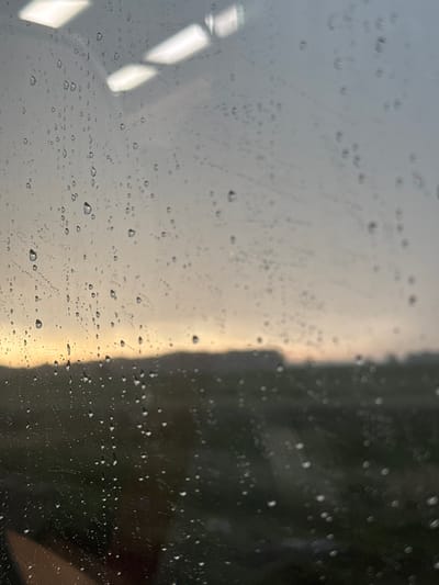 The image is a close-up view through a window with raindrops. The foreground is dominated by water droplets of various sizes on the glass, some appearing clear and spherical, others elongated and streaky, indicating movement. Beyond the window, the scene is out of focus and appears to be an outdoor environment with a muted color palette. There is a visible horizon line, suggesting a landscape. The light in the background is diffuse, with a soft orange and yellow glow near the horizon, possibly indicating sunrise or sunset. Above the horizon, the sky is a hazy gray.
This image was captured through a window at the location of Ul. Sowia in Kotowo, Poland. The scene depicts a rainy or recently rained-on environment viewed from an interior perspective. No people, specific objects, or actions are discernible beyond the raindrops and the blurred outdoor landscape. The lighting suggests a low-light condition.