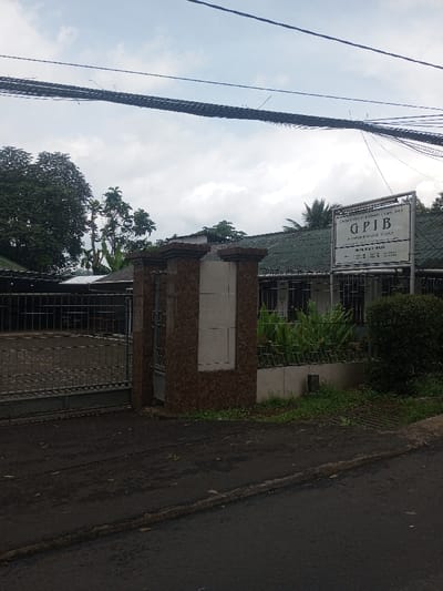 The image displays an outdoor, street-level perspective of a gated property and a building, ostensibly located in Chori, Nigeria, under an overcast sky. In the foreground, a dark, uneven paved surface, likely a roadway or driveway entrance, occupies the bottom of the frame. A large, dark grey metal gate with a grid design is visible on the left, flanked by two substantial pillars adorned with reddish-brown patterned tiles and integrated lighter beige panels. A smaller pedestrian gate is positioned to the right of the main gate within the pillar structure.
Beyond the gate, a paved area leads towards a building with a dark green tiled roof. The building's lower facade is partially obscured by a low metal fence and dense green foliage. Windows, protected by dark metal bars, are visible behind the plants. To the right of the entrance pillars, a prominent rectangular white sign with a dark frame is mounted. Legible black text on the sign reads: "GEREJA PROTESTAN DI INDONESIA BAGIAN BARAT", "GPIB", "JEMAAT BUMI SERPONG DAMAI", and "SEKTOR II".
Dense green trees and bushes are visible behind the property on the left and also contribute to the greenery surrounding the building on the right, with a manicured hedge present on the far right. Multiple dark electrical or communication cables span horizontally across the upper portion of the frame against the cloudy sky. No individuals, animals, or vehicles are present in the scene.