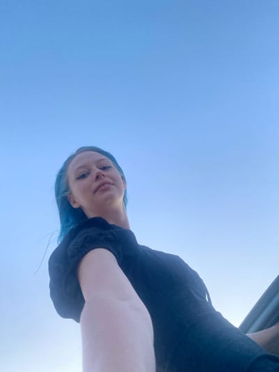 A low-angle, wide shot captures a young woman with bright blue hair against a clear blue sky. She is wearing a black top and is positioned in the foreground, looking upwards at the camera. Her right arm extends towards the camera, partially obscuring her face. The background is entirely the sky, indicating an outdoor setting. The image appears to have been taken in Milyanfan, Kyrgyzstan.