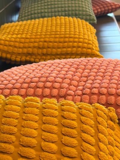 The image shows a close-up, angled view of several textured pillows stacked on top of each other. The pillows have a raised, bumpy pattern and are in various colors, including green, yellow, and coral. The lighting is soft, and the background is out of focus, suggesting an indoor setting. There are no people or specific landmarks visible in the image to indicate a location in Malmö-Sturup, Sweden.