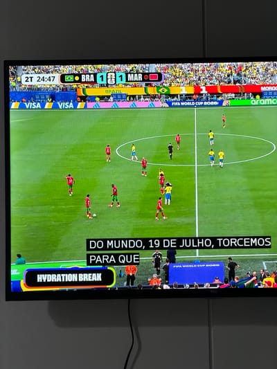 The image shows a soccer match broadcast on a television screen. The score is tied 1-1 between BRA and MAR. The time is 24:47 in the second half (2T). The text on the screen also includes "FIFA WORLD CUP 2026" and "HYDRATION BREAK". There are several players on the field, wearing red and yellow jerseys. The background shows a stadium filled with spectators and various advertisements. The broadcast is likely from a sporting event in João Pessoa, Brazil, given the prominence of Brazilian flags and the mention of "BRA".