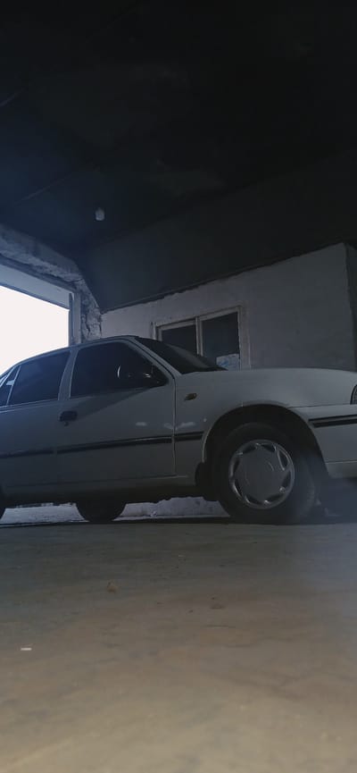 The media file displays a side view of a white van parked in a narrow alley. The van is positioned vertically within the frame. The alleyway is defined by a light-colored wall on the left and a darker building on the right, which has a window. The van is the primary subject, with its front wheel and part of the driver's side visible. The scene suggests an urban or semi-urban environment, likely in a densely built area given the narrowness of the passage. The lighting appears somewhat dim, possibly indicating early morning, late afternoon, or overcast weather conditions. The overall atmosphere is neutral, with no specific emotions or events clearly depicted. The image is taken from a low angle, looking slightly upwards towards the sky visible at the top of the frame. There is no visible text in the image. The location context is Kasansay, Uzbekistan.