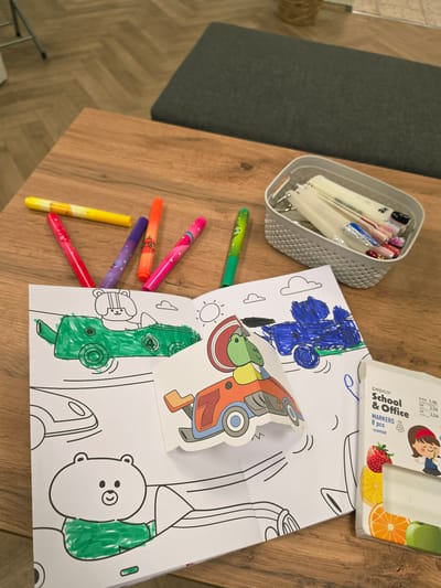 An overhead shot captures a creative activity unfolding on a warm-toned wooden table. The primary focus is a collection of coloring sheets featuring cartoon vehicles like a car, an airplane, and other whimsical figures including a bear. One airplane cutout, fully colored in shades of red, orange, green, and blue, is placed atop the main coloring sheet. Parts of the other illustrations are also colored; a car is filled in blue, a ground vehicle is green, and a bear's ear is green.

Scattered across the table are several brightly colored markers in shades of pink, orange, purple, and yellow, suggesting ongoing use. Another green marker lies apart from the cluster. In the upper left corner, a gray, textured container holds an assortment of nail sample sticks, some with intricate designs and colors, with faint text "VG" visible on one.

Adjacent to the coloring pages in the top right, a partially visible white box for "pepco School & Office MARKERS" is present. The box indicates "8 pcs -scented" and lists price information: "EUR/€ 1,30", "KM 2,50", and "лв. 2,54". The scene suggests an indoor setting, possibly a home or a leisure area in Mladá Boleslav, Czechia, characterized by the wooden table and a dark gray fabric-covered surface visible on the left.