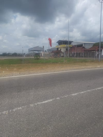 The image shows an airport control tower and buildings, a windsock, and a chain-link fence. The sky is cloudy and the ground is covered in grass and asphalt. The scene is a typical airport environment. The location is given as Glokaron, Indonesia. There are no people visible in the image. The focus is on the infrastructure of the airport. The image is taken from a roadside perspective, suggesting the viewer is on a road adjacent to the airport.