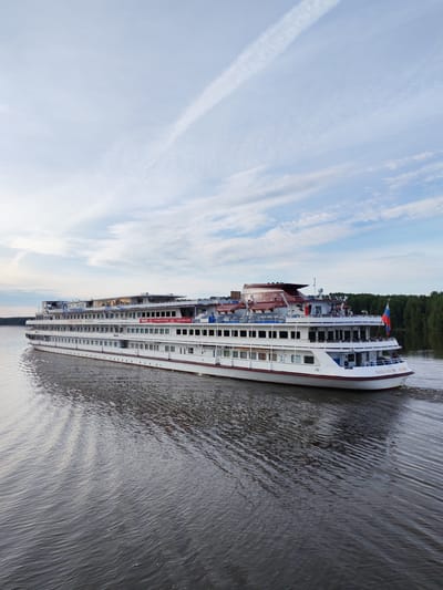 A large white river cruise ship, the "Konstantin Fedin," sails on a wide, calm river. The ship has multiple decks, each lined with numerous windows. A Russian flag is visible at the stern. The riverbanks are lined with a dense forest of green trees. The sky above is a pale blue with scattered white clouds and a visible contrail from an aircraft. The water reflects the sky and the ship, creating ripples and patterns. The scene appears to be set in the early evening or late afternoon, as indicated by the soft light. The image was taken from an elevated vantage point, possibly another boat or a bridge, looking down on the river and the ship. The location is identified as Krapivino, Russia.