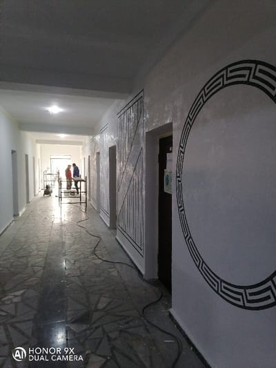 This image depicts a long, well-lit interior hallway in Samarkand, Uzbekistan, appearing to be under renovation or recently completed decorative work. The hallway features a dark-colored tiled floor with an irregular, mosaic-like pattern. The walls are light-colored; the right wall displays geometric decorative patterns in black, including a large, incomplete circular Greek key design and segmented rectangular designs with vertical and diagonal lines. Multiple dark doorways are visible along both walls. The ceiling is plain and light-colored, fitted with overhead light fixtures.
In the distance, at the far end of the hallway, two individuals are present. One wears a red upper garment and dark trousers, while the other wears a blue upper garment and dark trousers. They stand near a metal structure, possibly a small scaffolding or work table. To the left of them, near the wall, an air compressor or similar equipment with a tank is visible. A dark electrical cable snakes along the floor on the right side of the hallway, extending from the foreground towards the background. Natural light appears to enter from the very end of the corridor.