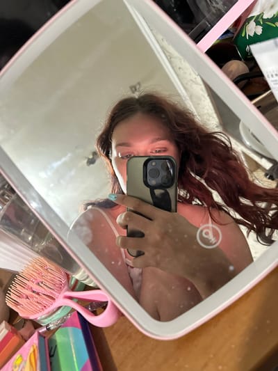 The image is a selfie taken in a mirror. The main subject is a young woman with long, dark red hair, holding a smartphone to take the photo. She is wearing a white tank top. Her face is partially visible, and her eyes are looking directly at the camera. She has a light makeup on, and her fingernails are painted a light blue-green color.
The mirror is rectangular with a pink frame. It appears to be a lighted vanity mirror, as a circular icon resembling a light bulb with a keyhole is visible on its surface, suggesting it might have a built-in light. The background reflected in the mirror shows a cluttered surface with various items. These include what appear to be makeup products, bottles, and a pink hairbrush with a distinctive design. The surface behind the mirror seems to be a wooden tabletop.
The scene appears to be indoors, likely in a personal space such as a bedroom or a vanity area. The lighting suggests it could be daytime, given the natural light reflection in the mirror, but the presence of the lighted mirror itself indicates an environment where artificial light is also available. There is no clear indication of the weather or specific time of day beyond the general indoor setting. The overall mood seems casual and self-focused, typical of a selfie. The visible text on items in the background is not legible. There are no specific cues pointing to Bishkek, Kyrgyzstan, in the image itself.