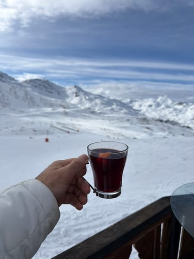 A right human hand, partially clad in a white insulated jacket, holds a clear glass mug containing a dark red liquid with an orange slice garnish. The mug is secured by a distinctive black metal handle extending from the rim to a base ring. The hand and mug are positioned over a dark wooden railing, with a segment of a circular glass tabletop visible on the far right. The background reveals an expansive, snow-covered alpine landscape, consistent with the ski domain of Saint-Martin-de-Belleville, France. Distant snow-capped mountains, visible ski slopes, and subtle red markers populate the midground. The sky above is partly cloudy with prominent horizontal cloud streaks. The overall scene depicts a cold, clear winter day in a high-altitude ski resort.
