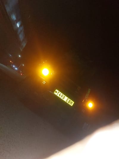 The scene is a nighttime view in Msata, Tanzania, focusing on the rear of a dark-colored vehicle. The vehicle displays two prominent, illuminated amber lights, likely tail lights or turn signals, and a rectangular yellow license plate bearing the registration 'T498 EBH'. A faint, smaller light is visible above and slightly to the left of the left amber light. The vehicle is positioned on a dark ground surface. The background is largely unlit, with a few distant artificial light sources visible in the upper left quadrant. A blurred, light-toned obstruction is present in the lower right foreground. No individuals are discernible.