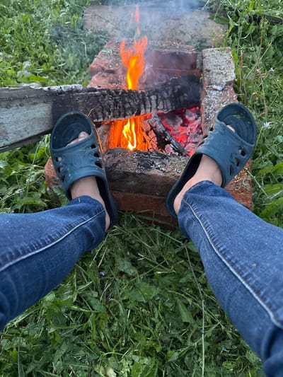 The image depicts a close-up view of a person's legs and feet, clad in blue jeans and dark blue slides, positioned in front of a bonfire. The bonfire is contained within a makeshift structure constructed from bricks and logs, situated on the ground amidst lush green grass. The flames are vibrant orange and yellow, casting a warm glow on the surrounding environment and the charred wood. The background shows a dense area of vegetation, suggesting an outdoor setting. The context provided places this scene in Imelevo, Russia.