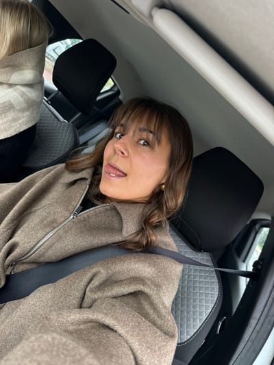 The scene depicts the interior of a vehicle, likely a car, in Mougins, France. A female individual, positioned in the foreground, directly faces the camera with a playful expression, her tongue extended. She has medium brown hair with bangs and wears a light brown, zippered jacket. A dark seatbelt crosses her torso and lap. A small, round, gold-colored earring is visible on her right ear. Her eyes are directed towards the camera. To the upper left, the back of a second individual's head with blonde hair is partially visible, wearing a light beige or cream-colored garment with a subtle pattern. The vehicle's interior features dark grey seats with a quilted texture, black headrests, and a light-colored overhead panel with a white grab handle. Through the side windows, a blurred green outdoor background is visible, with raindrops evident on the glass surfaces.