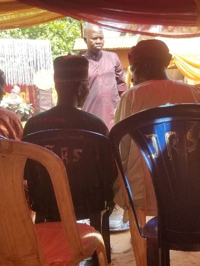 The image depicts an outdoor gathering in Ochadam, Nigeria, under a temporary canopy. In the foreground, two men are seated with their backs to the camera. The man on the left occupies a light beige plastic chair and wears a dark garment with "ERS" visible on the back, and a patterned dark cap. The man on the right sits in a dark blue plastic chair with gold "RS" lettering, wearing a light-colored garment and a red headwrap. A bald man with a short beard, dressed in a plum-colored collared shirt, stands in the midground, looking towards the viewer's left. Behind the seated figures, an electric fan is visible on the unpaved ground. The overhead canopy is fashioned from draped red and yellow fabrics. In the background, green foliage, a corrugated metal roof, and white decorative strips are present under bright daylight.