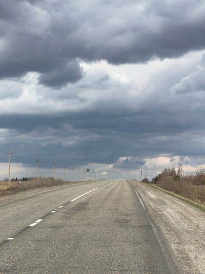 The image displays a long, straight asphalt road extending into the distance under a dramatic, cloudy sky. The road features a broken white lane marking in its center. The surface appears textured and slightly worn.
To the right of the road, a line of sparse, bare deciduous trees, possibly indicating late winter or early spring, separates the road from the distant horizon. Beyond these, a denser forest can be seen. A series of utility poles with power lines run parallel to the road on the right side, receding into the background. A circular road sign is faintly visible in the mid-ground, attached to one of the poles.
The sky is a prominent feature, dominated by a dynamic interplay of dark, heavy storm clouds and brighter, illuminated patches, suggesting an active weather front or recent rainfall. The scene is devoid of any human or animal activity, conveying a sense of solitude and vastness typical of a rural landscape.
It is daytime, but the heavy cloud cover casts a subdued light over the scene. The overall impression is one of quietude, perhaps hinting at changeable weather conditions in this rural setting, contextualized as Meshcheryaki, Russia. No discernible text is visible on the road signs or elsewhere in the image.