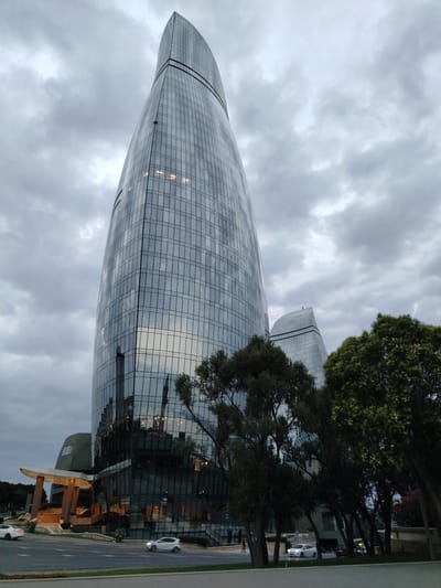 The image shows the Flame Towers in Sabail Raion, Azerbaijan, under a cloudy sky. The three flame-shaped skyscrapers are covered in a glass facade that reflects the sky and surrounding environment. In the foreground, a street is visible with two white cars and a dark-colored car driving on it. Trees with dense foliage are situated between the street and the towers. To the left of the central tower, a building with a covered entrance supported by columns illuminated with lights is present. The overall scene is captured from a low-angle perspective, emphasizing the height and grandeur of the Flame Towers.