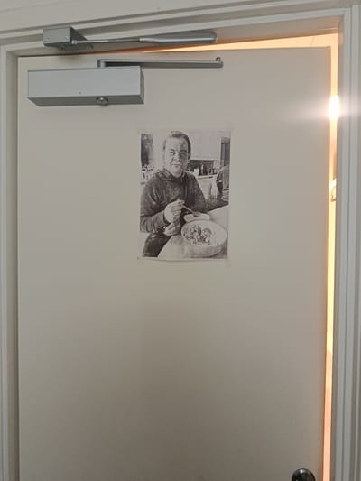 A black and white photograph of a man is taped to a closed, light-colored door. The man in the photograph is seated at a table, holding a spoon and eating from a bowl. He appears to be wearing a hooded sweatshirt. The door is equipped with a door closer mechanism at the top. To the right of the door, there is a narrow vertical gap through which bright light is emanating, suggesting an open space or hallway adjacent to the door. The scene is indoors. This image was taken in Vallberga, Sweden.