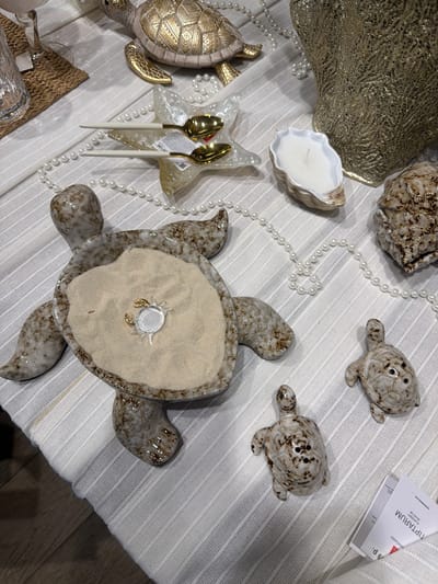 The image presents a still life arrangement on a white, horizontally striped surface. Several decorative elements are scattered across the scene. There's a large, textured golden object, possibly a platter, occupying the upper left corner. To the right of it, a beige-and-white shell-shaped object, likely a candle holder with a white candle inside, is visible. A string of white pearls meanders around these elements. Two small turtle figurines, with mottled brown patterns, are situated near the top of the frame. Further down, a larger, sand-filled turtle dish with a small, clear plastic crab toy in the center is placed. Two golden spoons with white handles lie beside a clear, starfish-shaped dish. To the lower left, there's a partially visible decorative turtle, adorned with gold patterns. An additional object made of woven material and a clear glass container with a metallic cap are situated at the very bottom left corner. A small white card with the text "TIPTARUM" is placed next to one of the turtle figurines. The setting suggests a decorative display. The mention of Novoalexandrovka, Russia is irrelevant, as the scene is contained within a still life arrangement and does not provide geographic context.