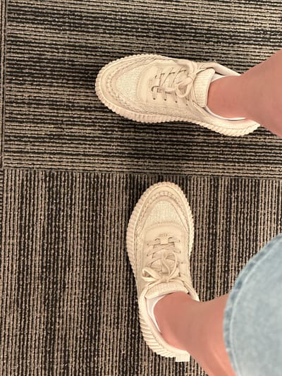 The image shows a top-down view of two feet wearing light-colored sneakers and white socks, standing on a carpeted floor. The carpet has a striped pattern with alternating dark and light grey horizontal lines in the upper half of the image, and dark and light grey vertical lines in the lower half. One foot is positioned in the upper right portion of the frame, with the sneaker pointing towards the upper left. The other foot is in the lower half of the frame, with the sneaker pointing towards the upper right. The scene is indoors. The location is specified as Lynnwood, United States.