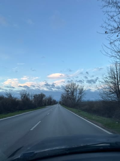 The image depicts a road in a rural area. The road is asphalt and has a single white line marking the center. Bare trees line both sides of the road. The sky is blue with scattered clouds. The photograph appears to be taken from inside a vehicle. The location in the city of Ohoden, Bulgaria is not evident from the image itself.