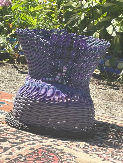 The image depicts a close-up, eye-level shot of a uniquely shaped, woven purple basket positioned outdoors on a patterned rug. The basket is the central subject, exhibiting a wide, flared base that narrows towards the middle and then widens again towards the top, creating an hourglass-like silhouette. The weaving is intricate, with alternating bands of darker and lighter purple creating a textured appearance. A thin, dark metal hoop extends from the top of the basket. Strands of small, pink and white beads are strung around the basket's midsection, adding a decorative element. The basket sits on a rug with a complex, multi-colored geometric and floral pattern in shades of red, beige, and black.

The environment surrounding the basket suggests an outdoor setting, possibly a garden or patio. In the background, lush green foliage from various plants, including some with broad leaves and others with smaller leaves and flowers, is visible. Sunlight filters through the leaves, casting dappled shadows. To the right of the basket, a small white daisy with a yellow center is in bloom. Behind the basket, a decorative element made of blue and white rounded objects, possibly bottles or stones, is partially visible, set against a darker ground. The ground itself appears to be a mixture of gravel and dirt. The lighting indicates it is daytime, with the sun casting a shadow of the basket onto the rug. There are no people or animals present in the image.
