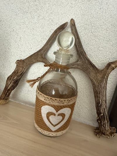 In Belo Pole, Bulgaria, on a light brown wooden shelf, a glass bottle with a clear stopper sits against a textured white wall. The bottle is decorated with a burlap wrap and a white heart cutout. Two deer antlers frame the bottle, one on each side, with their bases resting on the shelf. The left antler curves upwards and inwards, while the right antler curves upwards and outwards. The scene is lit from the front, casting subtle shadows.