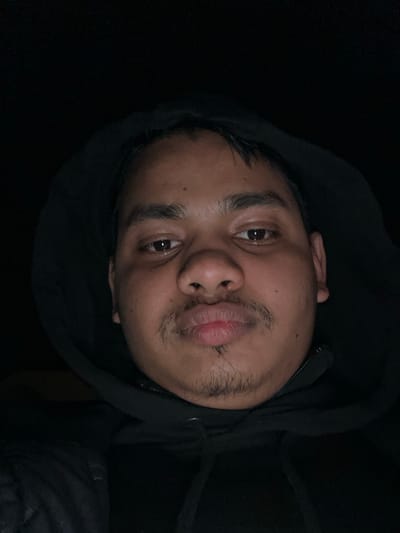 A close-up image depicts a single adult male, framed from the upper torso to the head, in Holtzheim, France. The male has a medium skin tone, dark brown eyes, a mustache, and a small goatee. He wears a black hooded sweatshirt with the hood pulled up, partially obscuring the top of his dark hair. The sweatshirt's zipper and drawstrings are visible below his chin. His lips are deliberately pursed, and he looks directly into the camera. The background is uniformly dark and devoid of any discernible features, indicating a low-light environment. The subject's face is illuminated by a direct light source from the front, consistent with a camera flash. No other individuals or objects are present.