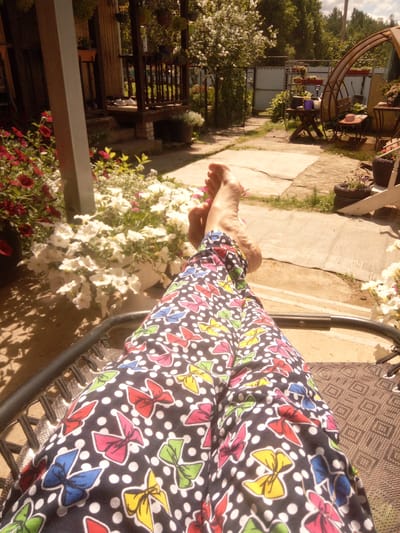 The image shows a person's legs, clad in black pants with a pattern of colorful bows and white polka dots, resting on a woven outdoor chair. The person's feet are visible, bare and angled upwards. The scene is set in a garden or yard. White petunias and red flowers are in bloom, and a portion of a wooden porch or structure is visible in the background, along with some greenery and a greenhouse-like structure. The ground is paved with concrete slabs. The lighting suggests it is daytime, with dappled sunlight and shadows. This scene is situated in a garden in the city of Pervomayskaya, Russia.