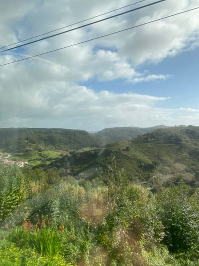 The image captures a panoramic view of a verdant, hilly landscape located in the rural vicinity of Mafra, Portugal. The upper third of the frame features a partly cloudy sky with patches of blue visible between scattered white cumulus clouds. Two dark, parallel overhead utility wires traverse the sky horizontally in the upper section. A faint, arc-shaped rainbow is discernible beneath the upper wire in the upper-left sky.
The mid-ground and background are dominated by rolling hills and valleys densely covered with dark green vegetation, including forests and cultivated fields. To the far left in a valley, a small cluster of white and light-colored buildings with reddish roofs, consistent with a village or hamlet, is visible amidst lighter green fields. A narrow, winding path or road is faintly visible traversing a hillside in the middle-right.
The immediate foreground is filled with abundant bright green foliage, including various bushes, grasses, and a cluster of red-orange flowers on the lower left. Sunlight creates several specular highlights and reflections across the foreground vegetation, indicating the presence of a reflective surface, possibly a window, through which the photograph was taken. No individuals or animate subjects are present in the scene, and no specific actions or interactions are depicted.
