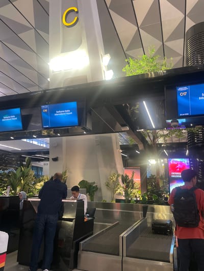 This media file depicts a scene at Soekarno-Hatta International Airport in Indonesia. The primary subjects are several people interacting at check-in counters. One individual, wearing a red shirt and a backpack, is standing near a screen displaying flight information. Another person, dressed in a dark jacket and jeans, is leaning over a counter, possibly interacting with airport staff. In the background, a person in a white shirt appears to be working at a counter.
The scene is an indoor airport environment with modern architecture. Large screens display flight details, including "Soekarno-Hatta International Airport" and gate numbers like "C16" and "C17," along with dates "22 Jun 2025" and "22 Jun 2026." Some screens also show "TIGA WARNA" and phone numbers. Lush green plants are strategically placed throughout the area, adding a natural element to the setting. The lighting suggests it could be daytime, with bright lights illuminating the space. The overall atmosphere appears to be a typical airport check-in process, with a focus on efficiency and passenger flow.