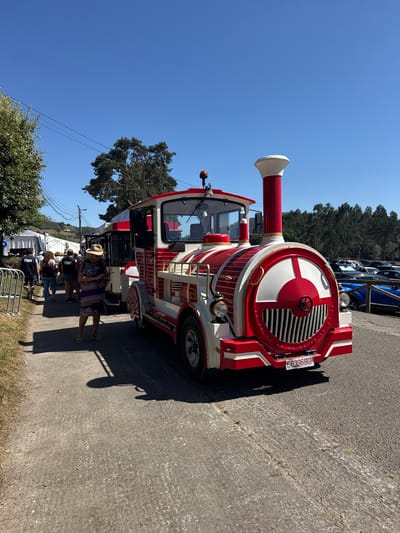The image shows a red and white replica train parked on an asphalt surface, likely outdoors. The train has a prominent front with a circular grille and two headlights. A person wearing a hat and a patterned dress stands to the left of the train, looking at their phone. Behind them, several individuals are visible, some wearing "STAFF" vests, indicating a possible event or organized gathering. The background reveals trees, a clear blue sky, and a parking lot filled with cars, including a blue sports car. The scene appears to be in a public area, possibly near a park or an outdoor venue. Based on the context provided, this scene is located in the city of Llanera, Spain.