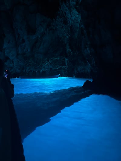 A boat floats on bright blue, illuminated water within a dark cave. The cave walls are rough and rocky, with a dark, shadowy texture. The water reflects the rocky ceiling. A person is visible on the left side of the frame, holding a phone and appearing to take a photo. The boat has people seated inside. The scene is illuminated by an unseen light source that causes the water to glow intensely blue. The location is the Blue Cave on the island of Biševo, near the town of Komiža, Croatia.