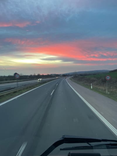 The image displays a forward-facing view from inside a vehicle, with the dashboard and two windshield wipers visible at the bottom of the frame. The scene is a multi-lane highway, located near Wallhausen, Germany, during twilight conditions.

The sky occupies the upper two-thirds of the image, characterized by horizontal bands of color: deep red and pink clouds in the mid-sky, transitioning to a pale yellow-orange glow at the horizon, and blue-grey with scattered clouds in the uppermost section.

The primary subject is an asphalt highway extending into the distance, appearing dark and intermittently reflective. It consists of at least two lanes in the direction of travel, separated by a dashed white line, and a solid white line marking the right edge of the roadway. A metal guardrail separates the primary highway from a visible parallel highway on the left. A circular speed limit sign, red-bordered with the number '100' in black, is positioned on the left side of the left highway. Multiple vehicles with illuminated headlights are observed traveling in the distance on both highways.

To the right of the main highway, a grassy shoulder and verge are present, alongside which a circular speed limit sign, red-bordered with the number '80' in black, is located. Beyond the verges, a low, gently rising landform with sparse vegetation is visible in the background. Deciduous trees with bare branches are present along the left side of the road.