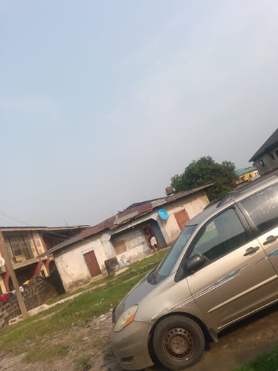 This image captures a daytime scene in Okere-Urhobo, Nigeria, featuring residential buildings and a parked vehicle under a hazy, light blue-grey sky.

In the foreground, a light gold-colored minivan, possibly a Toyota Sienna, is parked facing slightly right, occupying the lower right portion of the frame. Its side is adorned with blue stripe decals and appears splattered with a translucent liquid. The ground beneath and around the vehicle is unpaved, a mix of dirt, sparse green grass, and scattered debris.

The midground features a cluster of residential structures. To the left is a two-story building with a reddish-brown exterior and dark, possibly metal, grilles on its upper level. Clothes are visible hanging on a line or railing on the lower story. Adjacent to it, in the center, is a single-story building with a faded, light-colored facade and a rusty, corrugated metal roof. This structure has multiple brown wooden doors and at least two window openings, one appearing boarded up and another covered with a grid. A prominent blue satellite dish is mounted on its exterior wall. One individual, dressed in light clothing, is partially visible standing in the doorway of this central single-story building. A small, yellow water tank is visible on its roof.

In the background, additional residential buildings are discernible, including a darker, multi-story structure and a partially visible yellow-painted building, largely obscured by the dense green foliage of a mature tree. The sky occupies the upper half of the image, appearing clear but hazy.