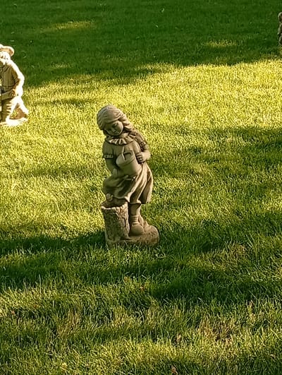 A statue of a young girl holding a watering can stands on a tree stump in a grassy yard. The statue is made of weathered stone and is detailed, with the girl wearing a dress and boots. Her head is tilted down, and she appears to be looking at the watering can. The statue is positioned in the center of the frame, with a lawn spreading out behind and to the sides. The grass is lush and green, with patches of sunlight dappling the ground. To the left of the main statue, another garden gnome-like statue of a person in work clothes is partially visible. The scene suggests a sunny day, likely in the late afternoon, given the long shadows cast by the sun. The overall mood is peaceful and idyllic, evoking a sense of childhood and nature. There is no visible text in the image. The location is specified as Pokrov, Ukraine.