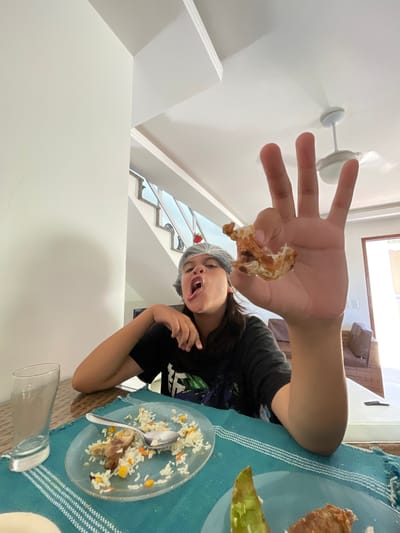 The image captures a candid, low-angle shot of a young person, likely a child or teenager, seated at a dining table indoors, possibly in a home in Cabo Frio, Brazil. The subject, with dark hair and wearing a black t-shirt and a grey hairnet or cap, is looking towards the camera with an expressive, wide-open mouth. Their right hand is held up close to the camera, prominently displaying a small piece of food, which appears to be a chicken bone with some meat still attached.

On the table in front of the person, there's a clear glass plate containing the remnants of a meal, consisting of rice mixed with corn and carrots, and another piece of meat or bone, along with a silver spoon. The table is covered with a blue and white patterned tablecloth. An empty, clear glass is also visible on the table.

The background features a bright, clean indoor setting with white walls. To the left, a white ceiling fan or light fixture is visible. To the right, a staircase with a dark railing ascends, and a window allows natural light into the space, indicating it is daytime. Partial views of white furniture or architectural elements are also present. The overall scene suggests a casual dining moment. No discernible text is visible in the image.