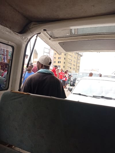 Inside a van, the back of a man wearing a brown shirt and a white baseball cap faces the camera. A light blue towel rests on his neck. Through the open back door, a busy street scene is visible. People, cars, and buildings form the backdrop. The van's interior shows a worn blue seat and a white ceiling. The scene suggests a ride in a crowded urban setting.