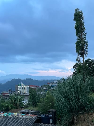 The image shows a view from a high point in Seetha Eliya, Sri Lanka. The sky is overcast with blue and gray clouds. Below the sky are rolling hills and valleys. Buildings and houses are visible in the foreground, with trees and other vegetation. The roofs are a combination of tile and corrugated metal. A mosque is visible among the buildings, distinguished by its white walls and green domes. A tall, thin tree is located on the left side of the sky.