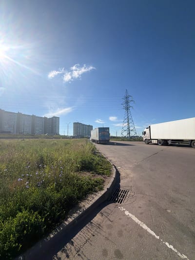 The image depicts an outdoor scene in the Settlement of sanatorium "Podmoskovye", Russia, under a clear blue sky with a bright sun and a few scattered clouds. In the foreground, a paved road curves to the right, with a grass verge and some low-lying green bushes and small purple flowers along its edge. A storm drain grate is visible on the road. In the middle ground, two large white trucks are driving on the road. The truck on the right is closer to the viewer and appears to be a semi-trailer truck. Behind the trucks, a tall, green lattice metal electricity pylon stands prominently. In the background, several large, multi-story apartment buildings are visible, suggesting an urban or suburban residential area. The overall impression is a bright, sunny day in a developed area with infrastructure and residential housing.