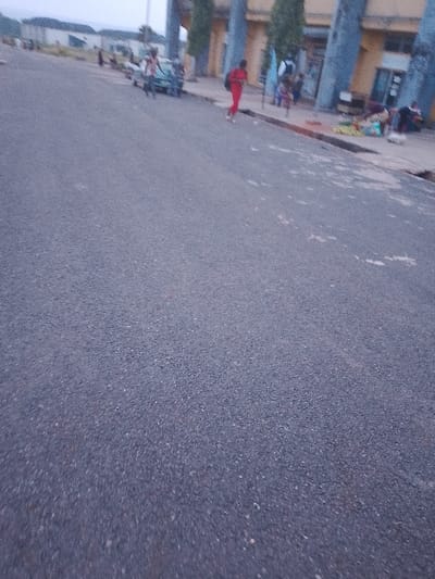 The image shows a street scene in Ogboko, Nigeria. The foreground is dominated by an asphalt road, while in the background there are buildings with large pillars and a few people. There is a person wearing a red jumpsuit walking along the sidewalk. Further back, there are several individuals gathered near the building, some appearing to be sitting or standing. A vehicle is also visible nearby. Some items that could be merchandise are visible near the building.