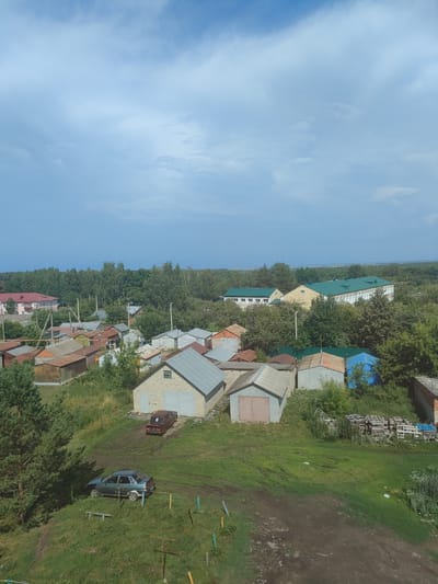 The image shows a collection of garages and residential buildings in what appears to be a rural or semi-rural area. The sky is overcast with a mix of blue and grey clouds. In the foreground, there are several small, single-story garages with various roofing materials, some made of corrugated metal and others with tiled roofs. A dark-colored sedan is parked on a dirt path next to a grassy slope. Further into the scene, more garages are visible, some constructed from brick and others with metal siding. A light-colored, brick building with a grey, pitched roof and double garage doors is prominent. In the middle ground, there are more residential-style buildings with red and green roofs, interspersed with trees. A line of trees forms a dense treeline in the background, separating the built-up area from what could be open fields or more distant vegetation. The overall impression is one of a community of garages and homes, likely in a suburban or village setting within Sovkhoz "Kommunar", Russia. There are no people visible in the image.