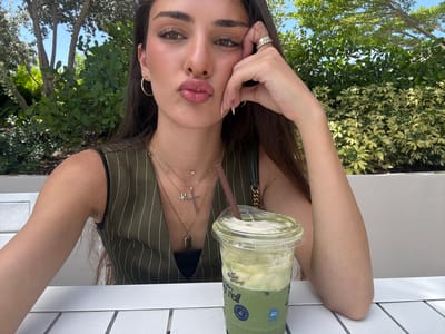 In Hallandale Beach, Florida, USA, a young woman with long, dark hair and makeup sits at a white table outdoors. She wears a green, pinstriped vest over a black top, adorned with several gold necklaces, including one with a cross pendant and another with a rectangular tag. A gold hoop earring dangles from her left ear, and she sports multiple rings on her right hand, one with a checkered pattern. Her right arm rests on her cheek, and her lips are puckered. In front of her is a clear plastic cup filled with a green beverage, likely a matcha latte, with a brown straw. The background features lush green foliage and trees under a bright blue sky.