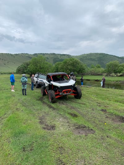 The image depicts an outdoor scene in a grassy, somewhat muddy field near a body of water, with rolling, tree-covered hills in the background under an overcast sky. It appears to be located in Yelantsy, Russia.

In the foreground, a white and orange off-road vehicle (side-by-side) is parked. To its left, a white SUV with a roof rack is partially stuck in the mud. Several people are present:

*   A person in a blue jacket and green shorts stands to the left of the vehicles.
*   A person wearing a helmet and a green and grey outfit stands near the side-by-side.
*   A person in a grey jacket and blue jeans stands near the SUV.
*   A person in a brown outfit is standing on the bank of the water to the right, holding a light blue item.
*   Another person in dark clothing and blue pants is standing near the water's edge.
*   A person wearing a helmet and camouflage jacket is standing between the two vehicles.

The environment suggests off-road recreational activity. The ground shows tire tracks and mud, indicative of recent vehicle movement in a wet area. The water appears to be a river or stream. The overall impression is one of a group engaged in outdoor adventure or recovery operations.