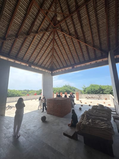 The image depicts an open-air structure with a high, conical thatched roof and exposed wooden beams, supported by concrete columns. The floor is a light-grey concrete. This structure is located in San Isidro Llano Grande, Mexico, with an open view to an outdoor terrace and a horizon of dense green foliage under a clear blue sky.
Approximately eight to ten individuals are present. In the foreground left, a person in a white jumpsuit stands with their back to the viewer, appearing to observe or photograph the scene. In the mid-ground right, another person with dark hair sits on a low wooden bench. Several other individuals are scattered in the mid-ground and background, standing in small groups, observing, or moving around objects.
Key objects within the covered area include a large, centrally located rectangular stack of reddish-brown bricks, approximately 1.5 meters tall, and to its right, a long, low wooden table covered with numerous bundles of light brown fibrous material, possibly burlap or dried straw. Several dark, spherical objects, resembling coconuts, are scattered on the concrete floor.
On the outdoor terrace beyond the concrete structure, multiple vertical stacks of reddish-brown bricks are arranged in rows. Scattered on this outdoor ground are several small, round, reddish-brown objects, likely terracotta, and a collection of larger, natural-colored items, possibly pottery. Towards the far left of the outdoor area, three distinct large red heart-shaped objects are visible against the green foliage.