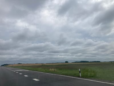 The image depicts a rural landscape in Лугаваслабадскі сельскі Савет, Belarus. A paved road occupies the foreground, with white directional markings and a solid white line delineating the lanes. To the right of the road, a striped marker post is visible. Beyond the road, a field of green and dry grass stretches into the distance, transitioning into a line of trees and vegetation along the horizon. The sky is overcast with heavy, grey clouds. The overall impression is one of a cloudy day in a rural setting.