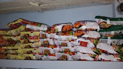 A large quantity of instant noodle packages are stacked on a shelf in a room in Kecamatan Panjalu, Indonesia. The packages are arranged in three rows. The top row consists of four packages of "Indomie Mi Goreng" and two packages of "Indomie Soto Ayam" with a red and yellow color scheme. The middle row consists of eight packages of "Indomie Goreng" with a white and red color scheme. The bottom row consists of eleven packages of "Indomie Kuah Soto" with a green, white, and red color scheme.  The packages are arranged neatly and are all facing the same direction. There is an overhead light source, the ceiling is blue and white, and there are no people present. The image was taken from a low angle.