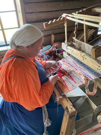 A person wearing an orange polka-dot blouse and a blue apron is weaving on a wooden loom in Boyarshchina, Russia. The person is wearing a white headscarf. The loom is made of wood and has threads of various colors woven into it. The person is holding a pair of scissors and appears to be cutting a piece of fabric. The background is a wooden wall and a window. The scene suggests a historical or cultural setting, possibly a museum or a traditional craft workshop.